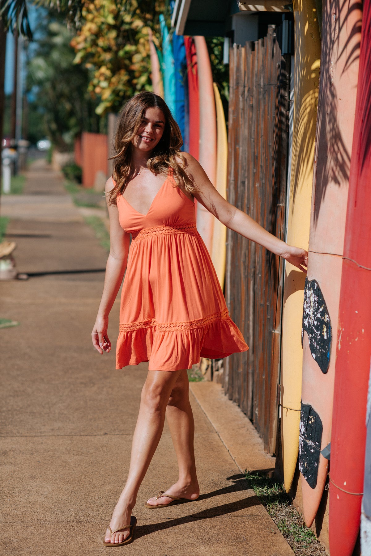 a burnette hair model posing on the street besides pink surf boards weaing a punch coral color mini dress from Koy Resort with on hand touching the surf board and matching the dress with brown flip flops
