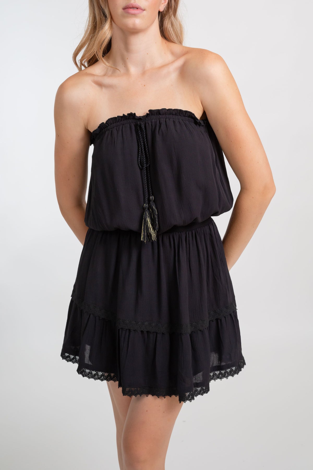 A blonde Caucasian model wearing a black color bandeau  crochet lace trim mini boho dress with metallic tassel trim, standing in a studio with her arms behind her back looking at the camera posing.