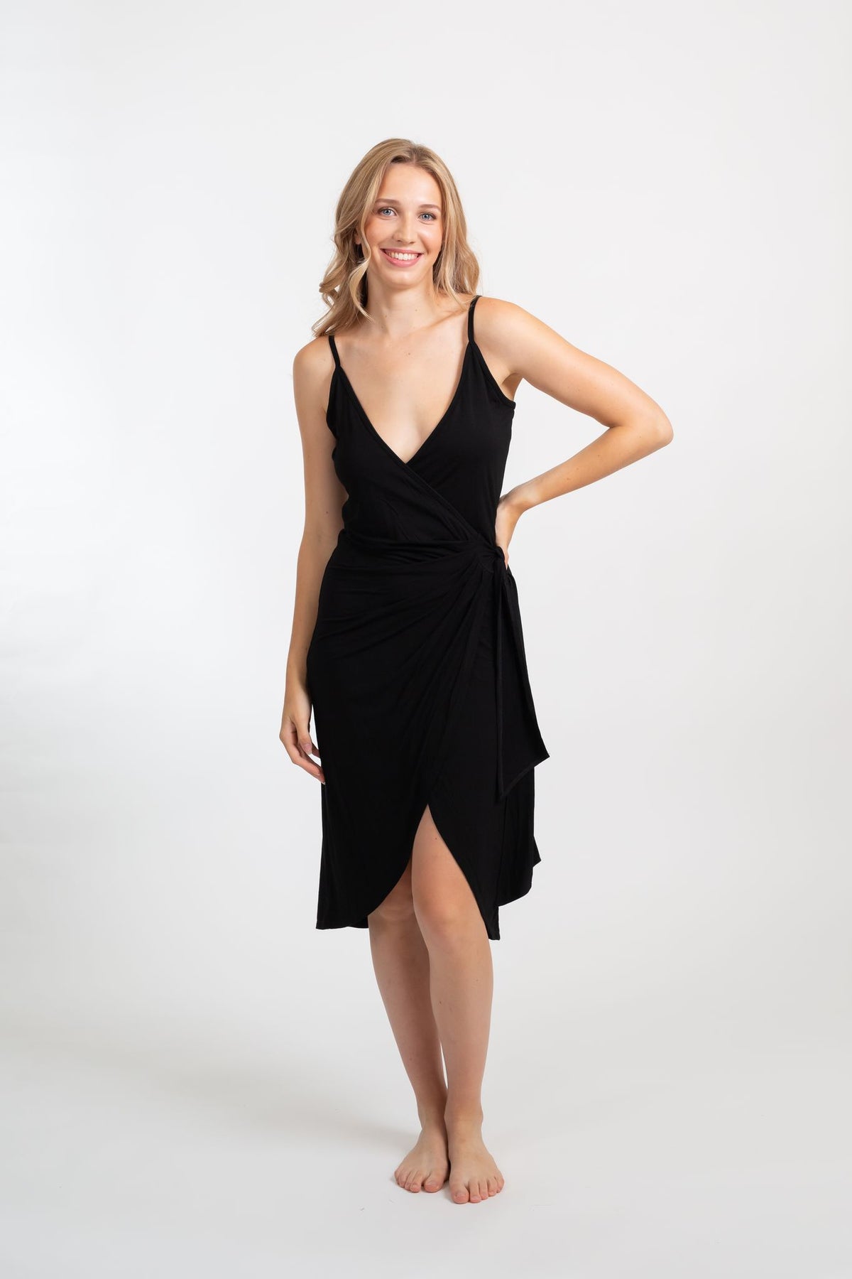the front of a blonde hair woman model wearing a strappy black dress that has a small cut in the middle with one hand on her hip