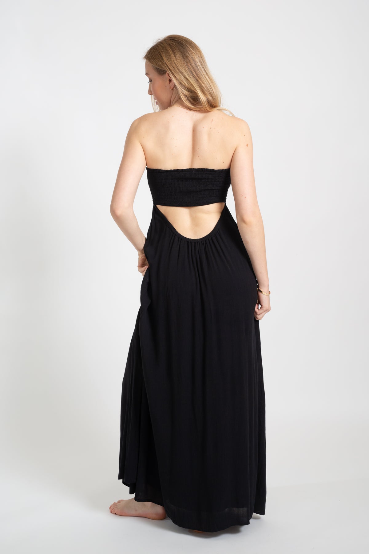 Blonde model wearing the black Miami maxi bandeau dress with the slit by Koy Resort Beachwear collection facing back. 