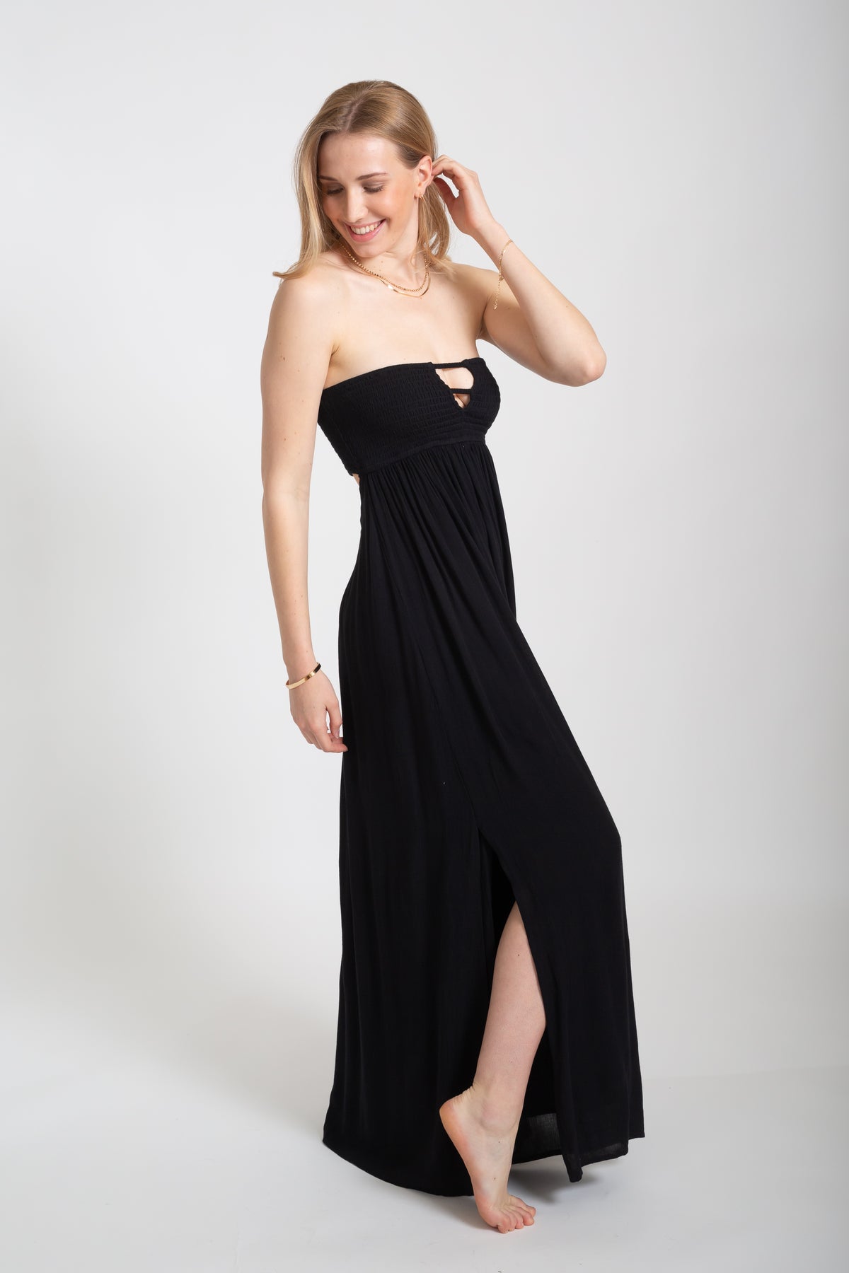 Blonde model wearing the black Miami maxi bandeau dress with the slit by Koy Resort Beachwear collection facing side.