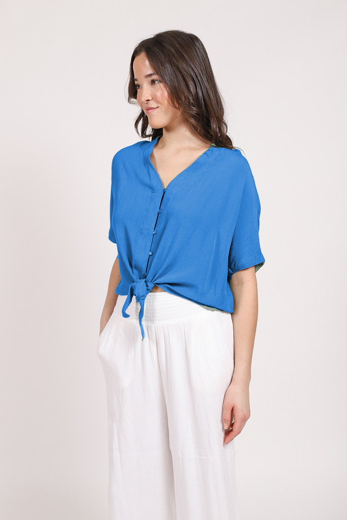 a dark hair female model looking to the side wearing a bright blue front tie button up shirt pairing with white pants