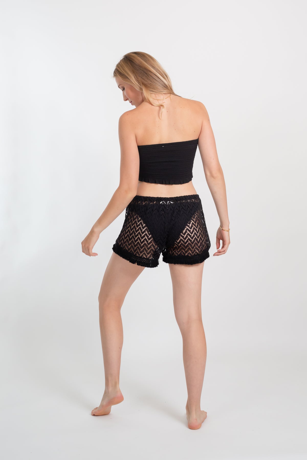 a back shot of a blonde hair female model wearing black bandeau top and black crochet lightweight mini beach cover up shorts from Koy Resort