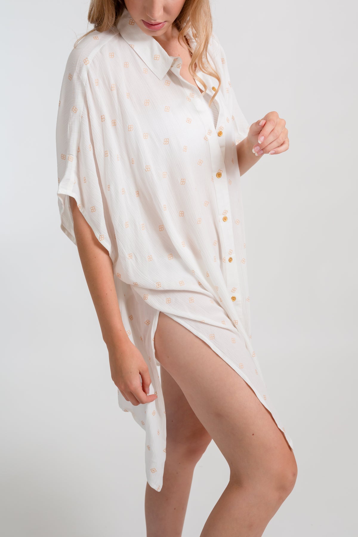 a zoomed in side photo a blonde hair female model wearing a buttoned up shirt dress with a side slit in the color white with gold dot print