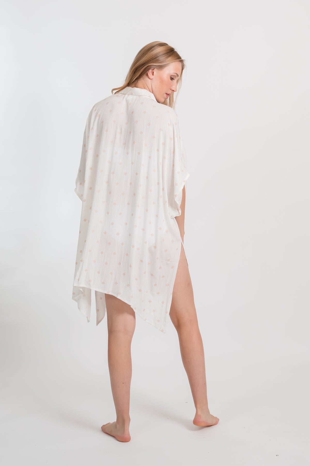 the back of a blonde hair female model looking down wearing a flowy big shirt dress in white with gold dot print