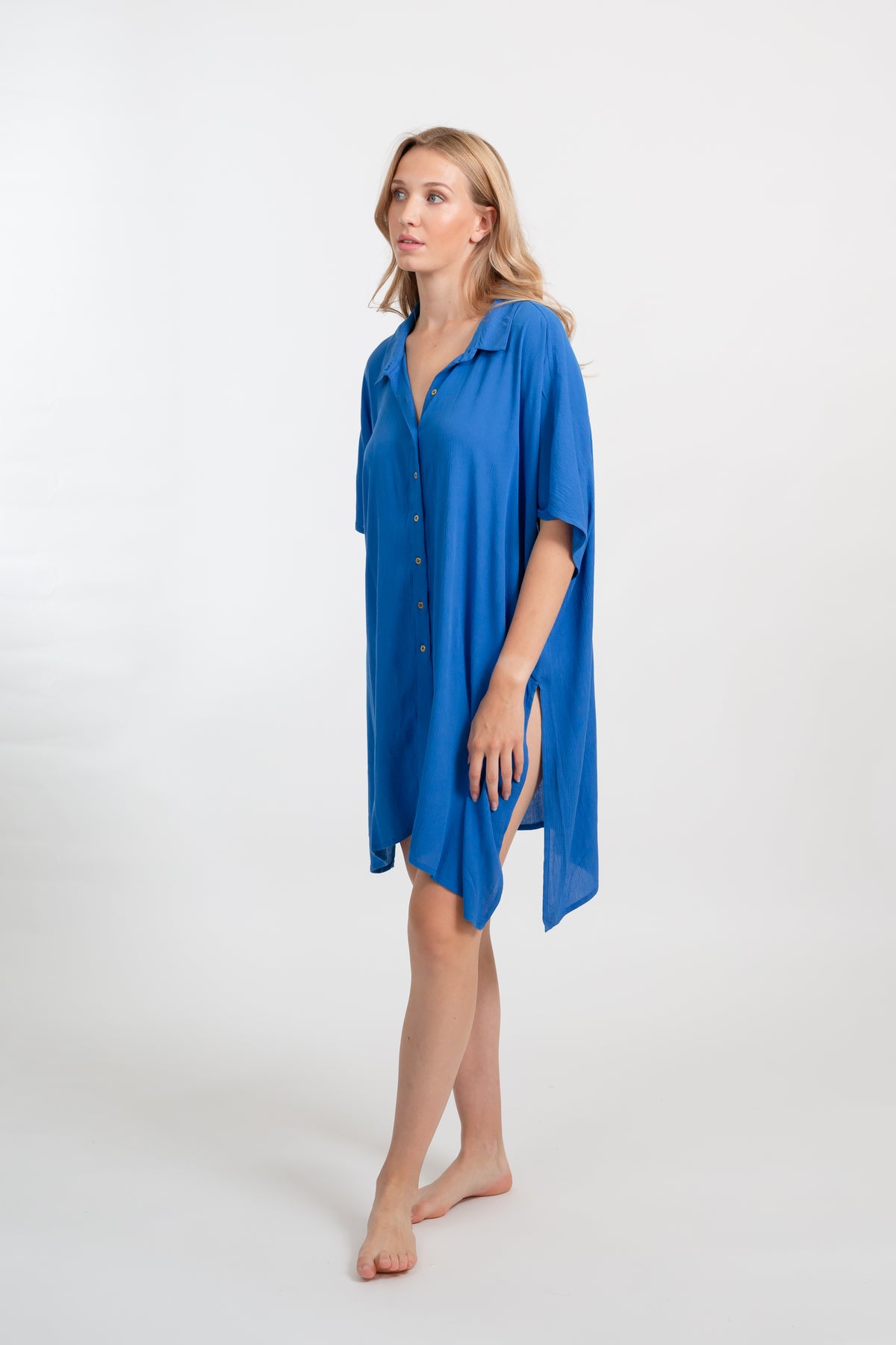 A blonde Caucasian model wearing a blue big shirt bathing suit cover up shirt dress, standing in a studio smiling.