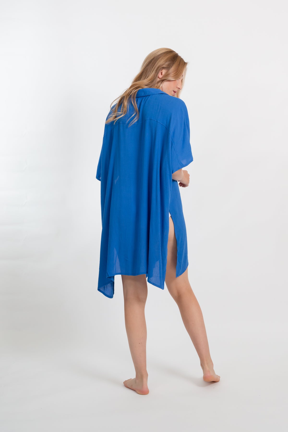 A blonde Caucasian model wearing a blue big shirt bathing suit cover up shirt dress, standing in a studio smiling and showing the back of the mini dress. 