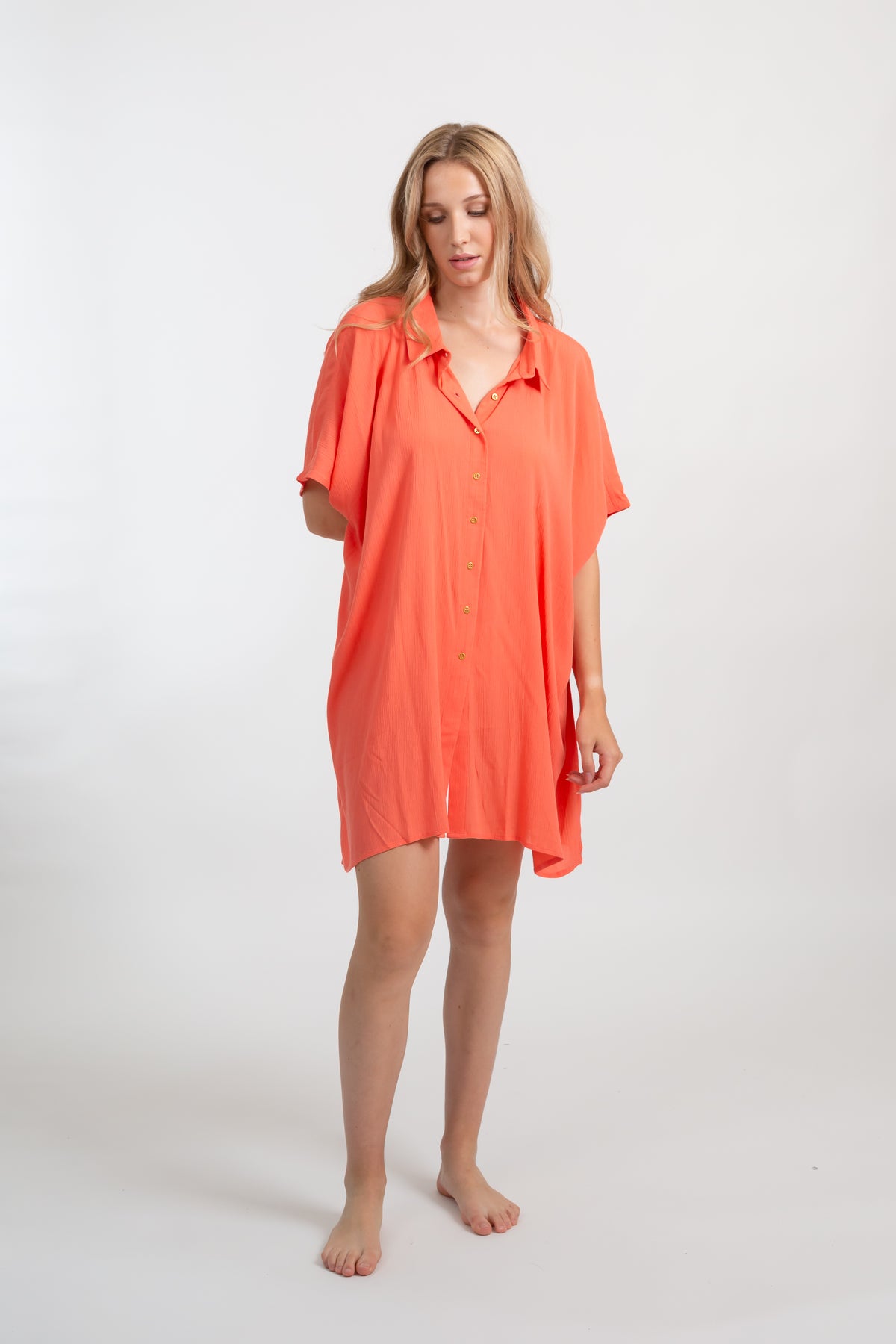 A blonde Caucasian model wearing a orange coral colored in "punch"  big shirt bathing suit cover up shirt dress, standing in a studio.