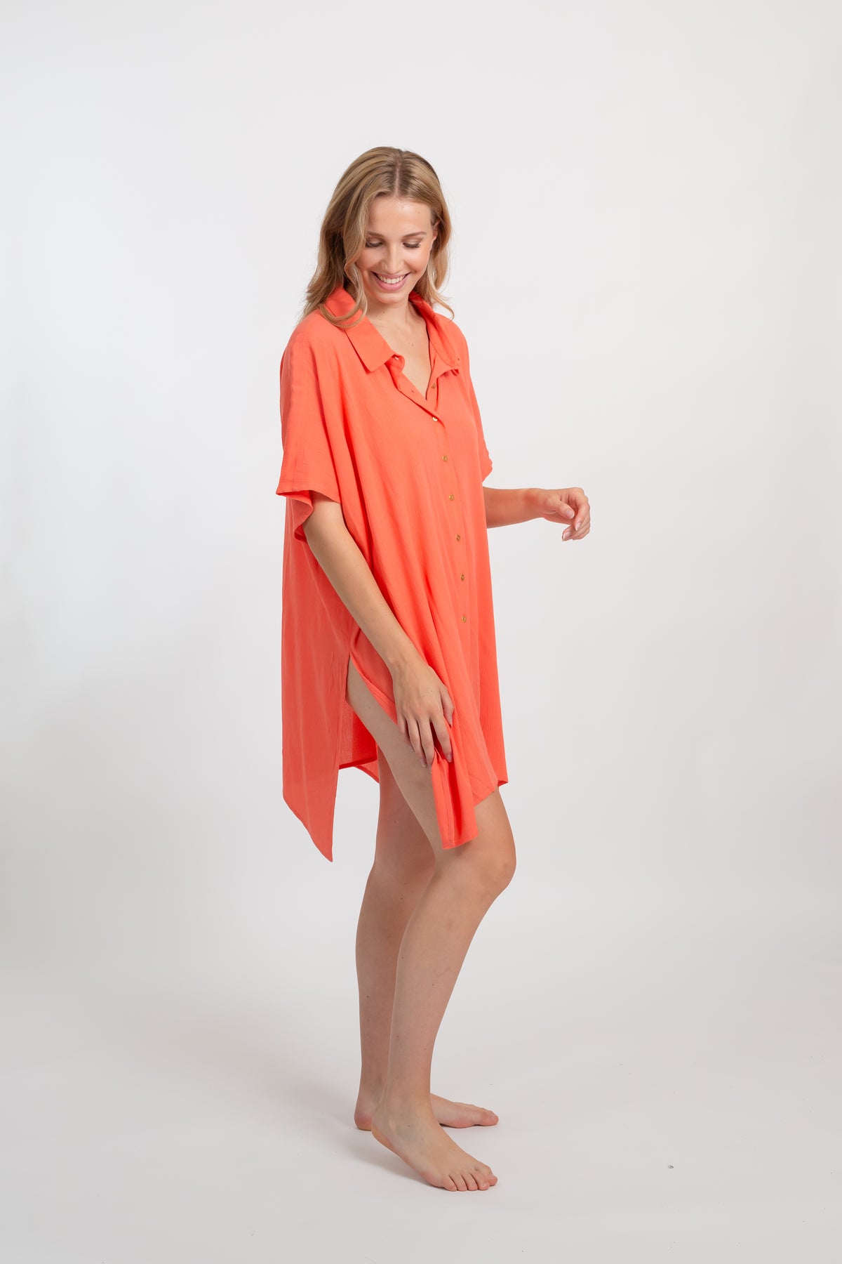 A blonde Caucasian model wearing a orange coral colored in "punch" big shirt bathing suit cover up shirt dress, standing in a studio, smiling and laughing. 