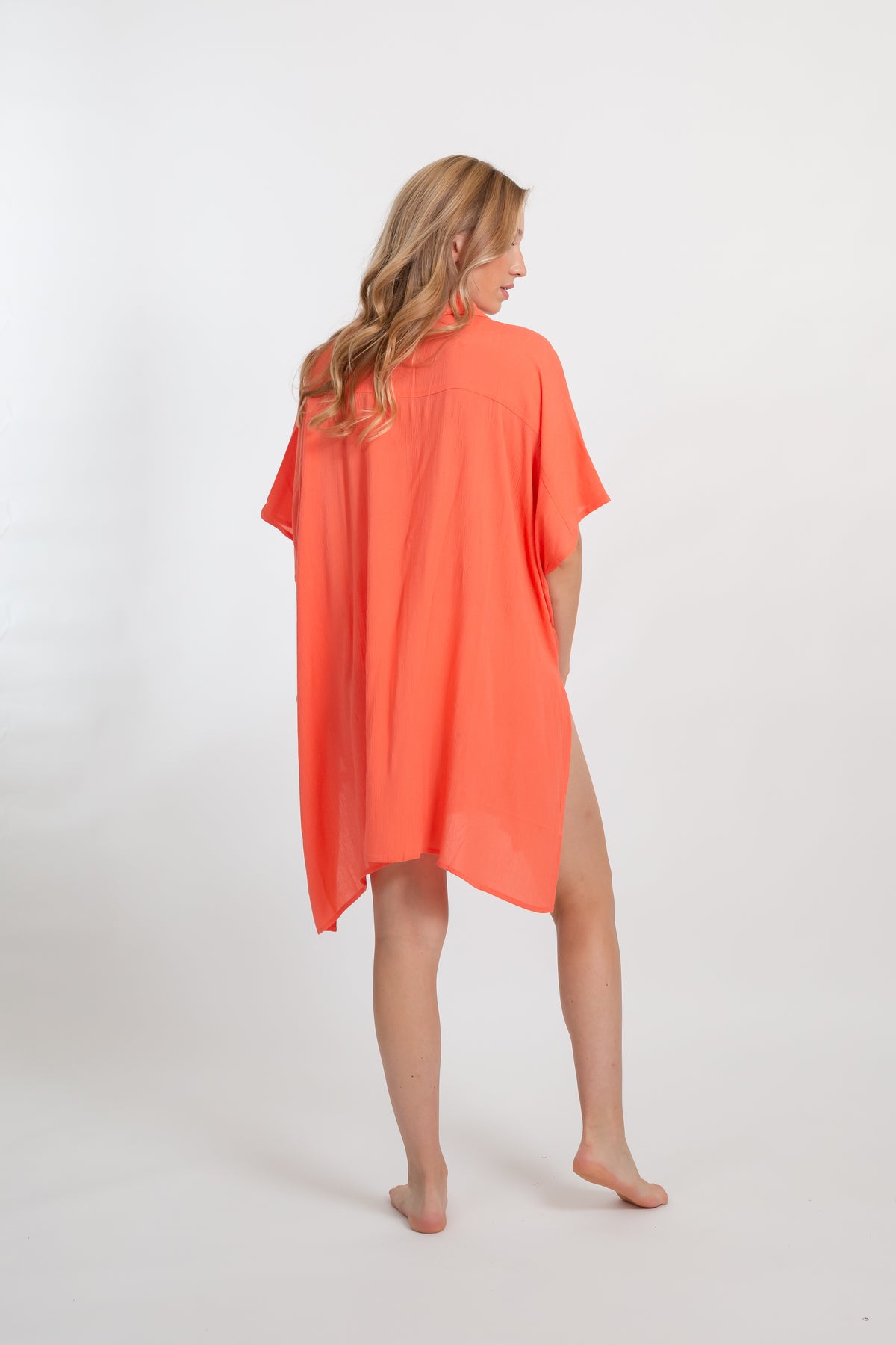 A blonde Caucasian model wearing a orange coral colored in "punch" big shirt bathing suit cover up shirt dress, standing in a studio, with her back facing towards and looking over her shoulder. 