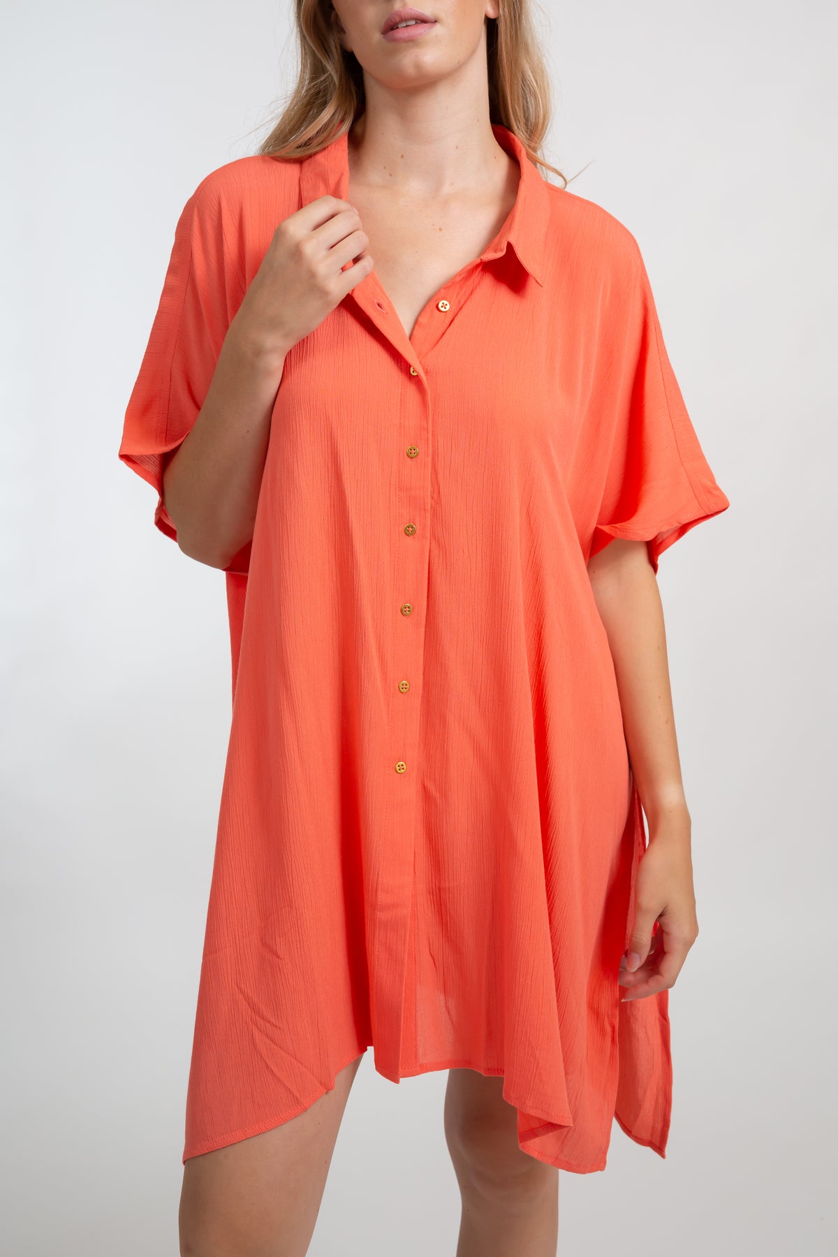 A blonde Caucasian model wearing a orange coral colored in "punch" big shirt bathing suit cover up shirt dress, standing in a studio.