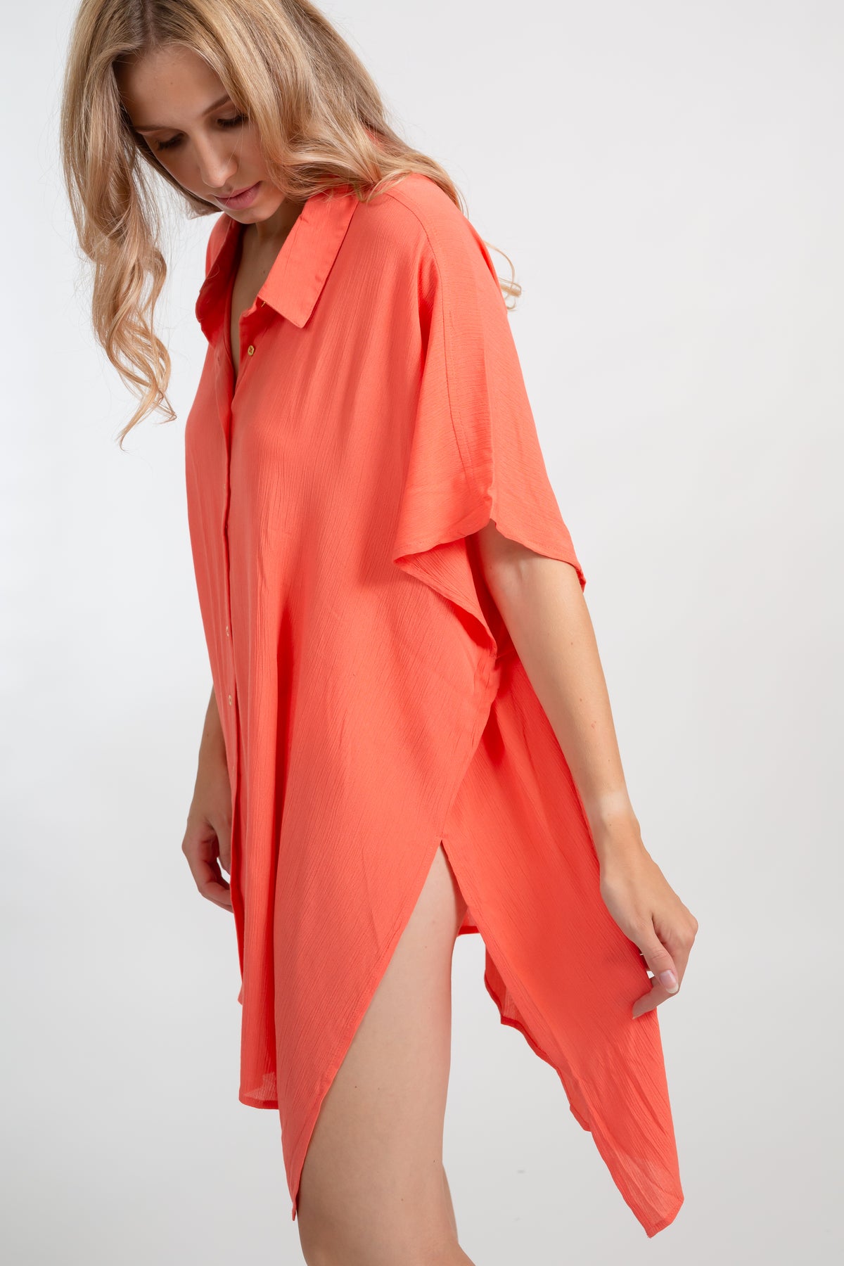 A blonde Caucasian model wearing a orange coral colored in "punch" big shirt bathing suit cover up shirt dress, standing in a studio, on the side pulling on the side of the dress to show the high slit design element. 