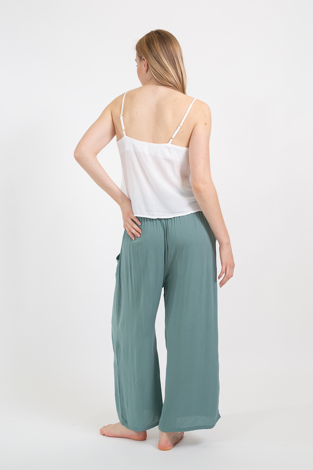 Blonde model wearing the white soft Miami Camisole top with adjustable straps and v-neckline facing back. Paired with crinkle rayon jade green Miami pants by Koy Resort Cruise summer beachwear collection.