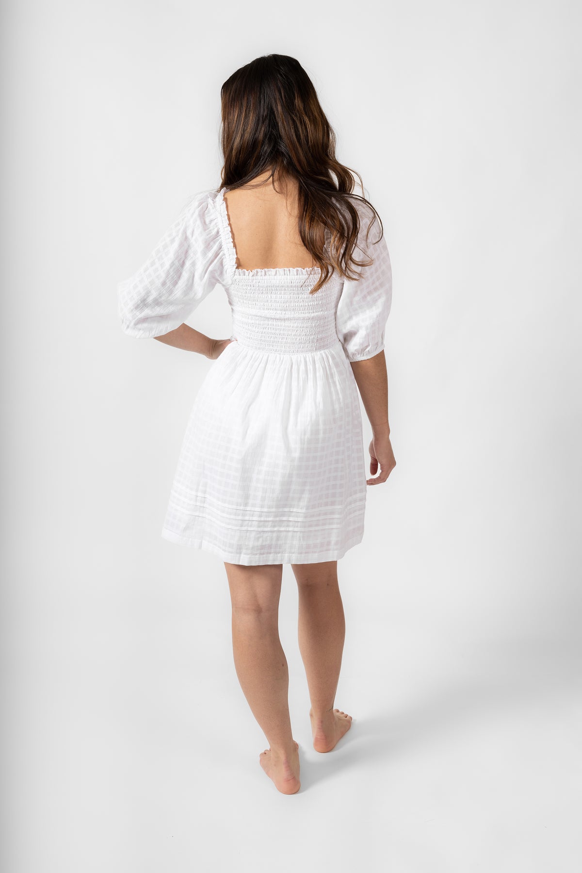 the back of a brunette hair model wearing a square back smocked short sleeve white dress from Koy Resort with one hand on her waist