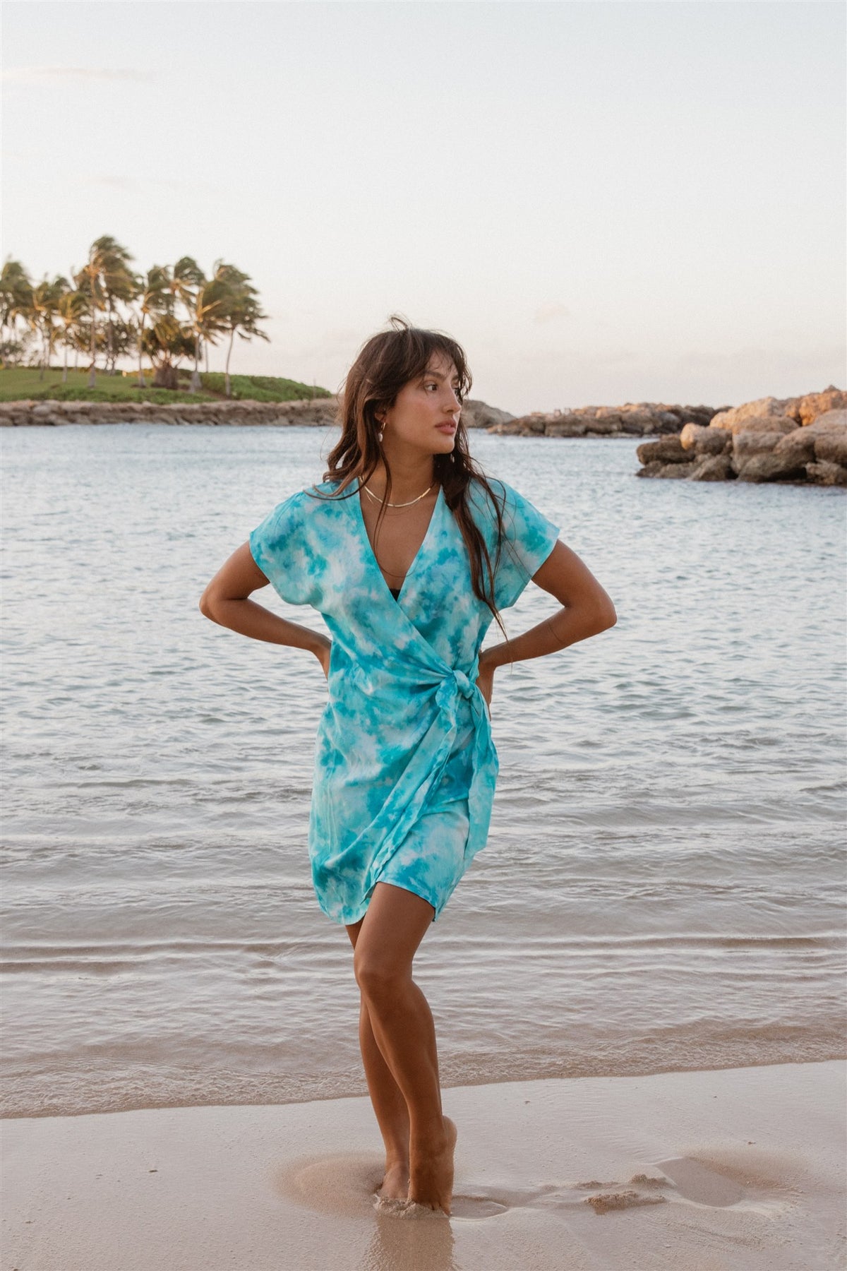 A women standing on the beach of Hawaii with a beautiful ocean view as the background. She has both arms on her waist and she is wearing a beautiful blue tie-dye mini wrap dress.