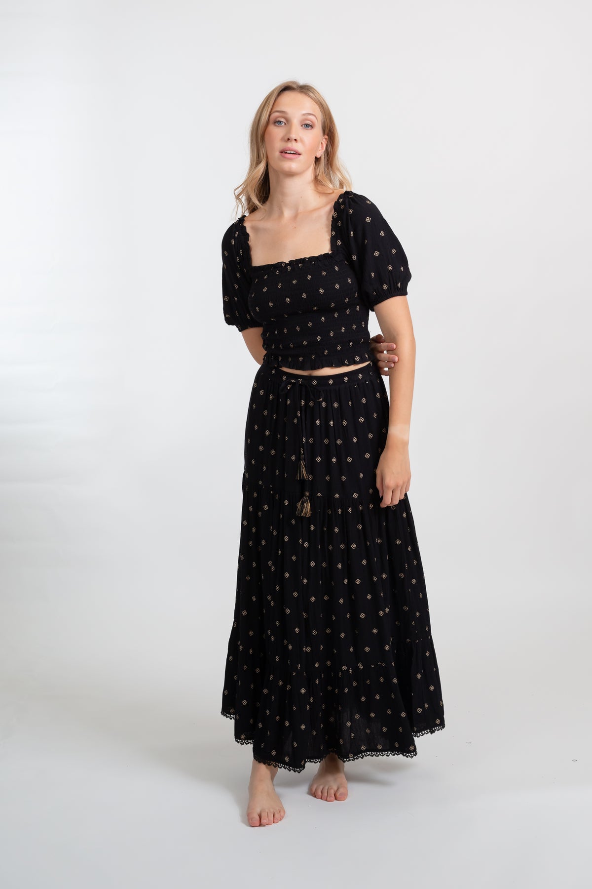 a front shot of a blonde hair woman wearing black with dot print matching smocked crop top and maxi skirt with one arm behind her back