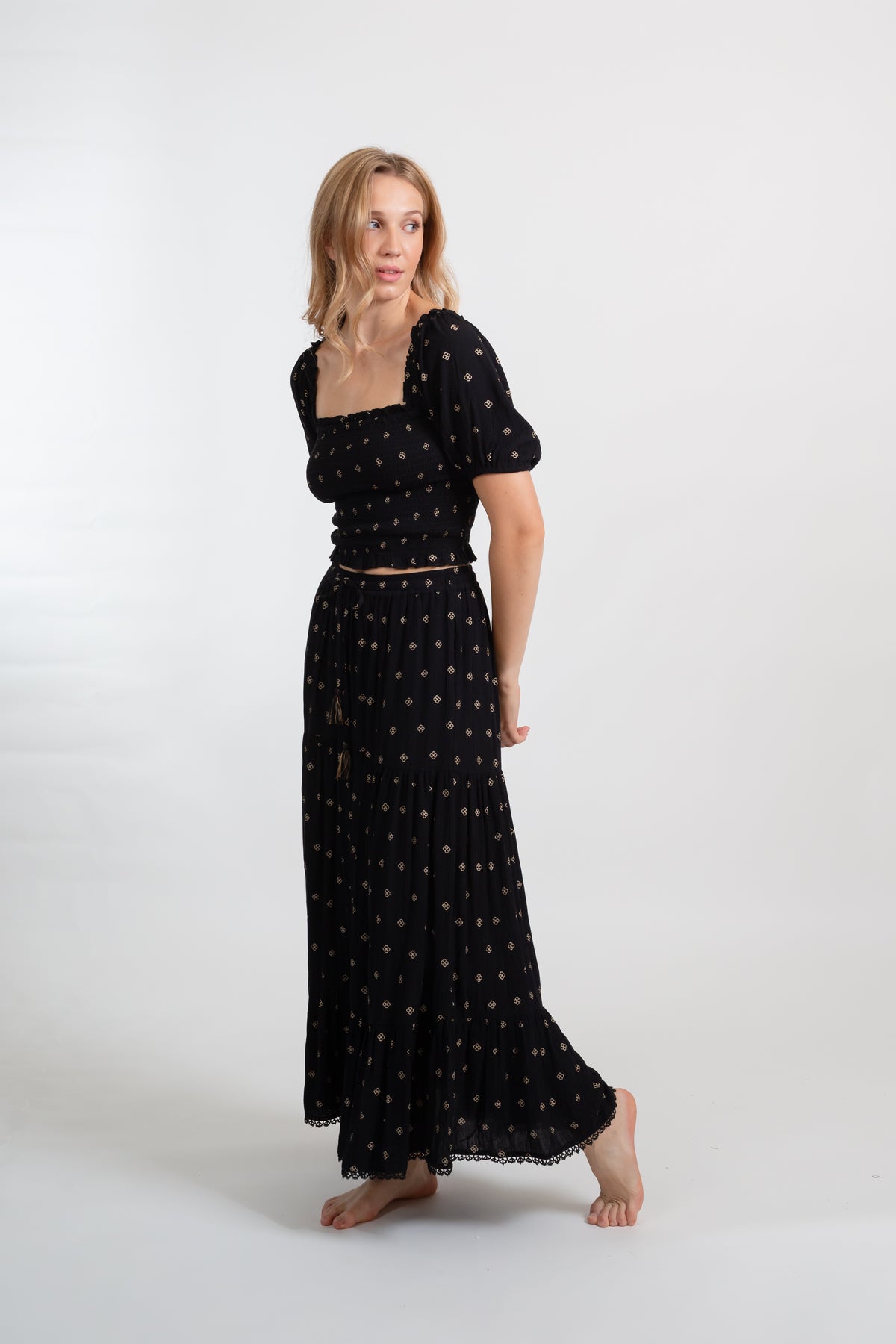 a blonde hair model wearing a black with gold dot print two piece smocked crop top and maxi skirt set from Koy Resort