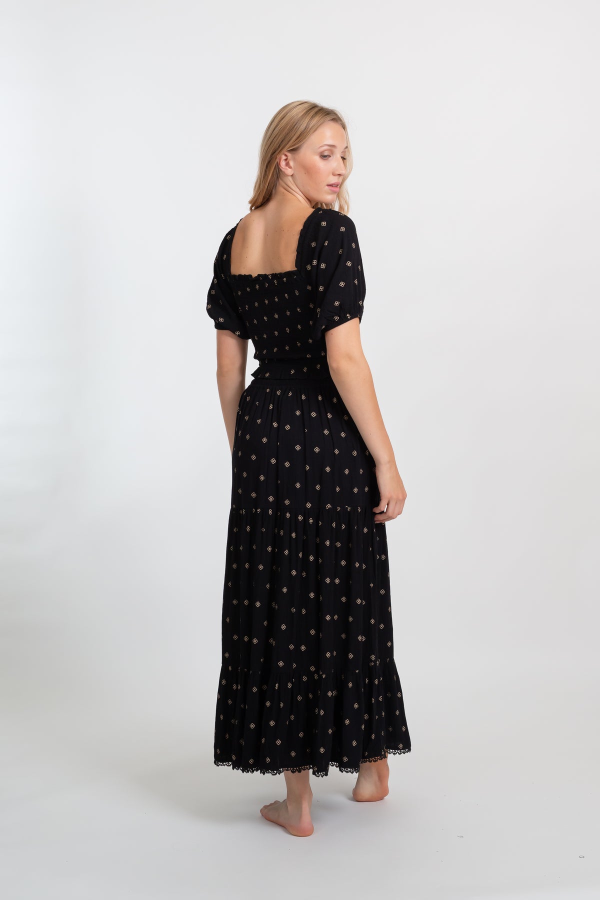 the back of a blonde hair woman model wearing a smocked square back black with dot print crop top and a black flowy black with dot print maxi skirt looking back