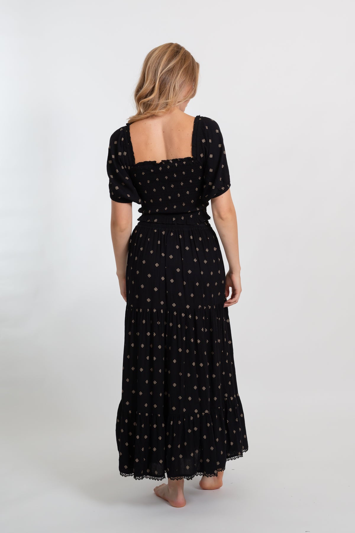 the back of a blonde hair woman model wearing a smocked square back black with dot print crop top and a black flowy black with dot print maxi skirt