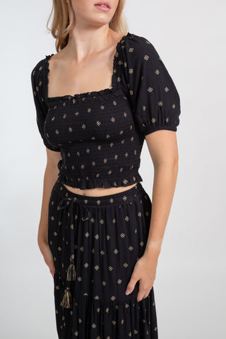 a zoomed in shot of a blonde hair woman model wearing matching black with gold dotted print smocked crop top and flowy dress