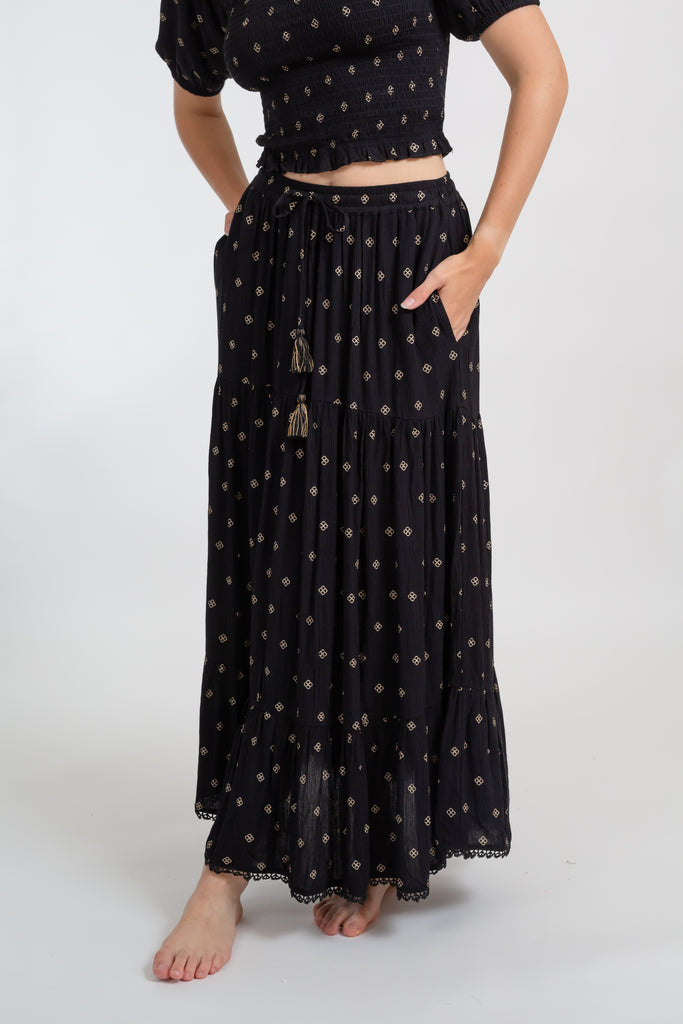 a zoomed in shot of a woman wearing a matching two piece black with gold dot print crop top and midi dress set with one hand in her dress pocket
