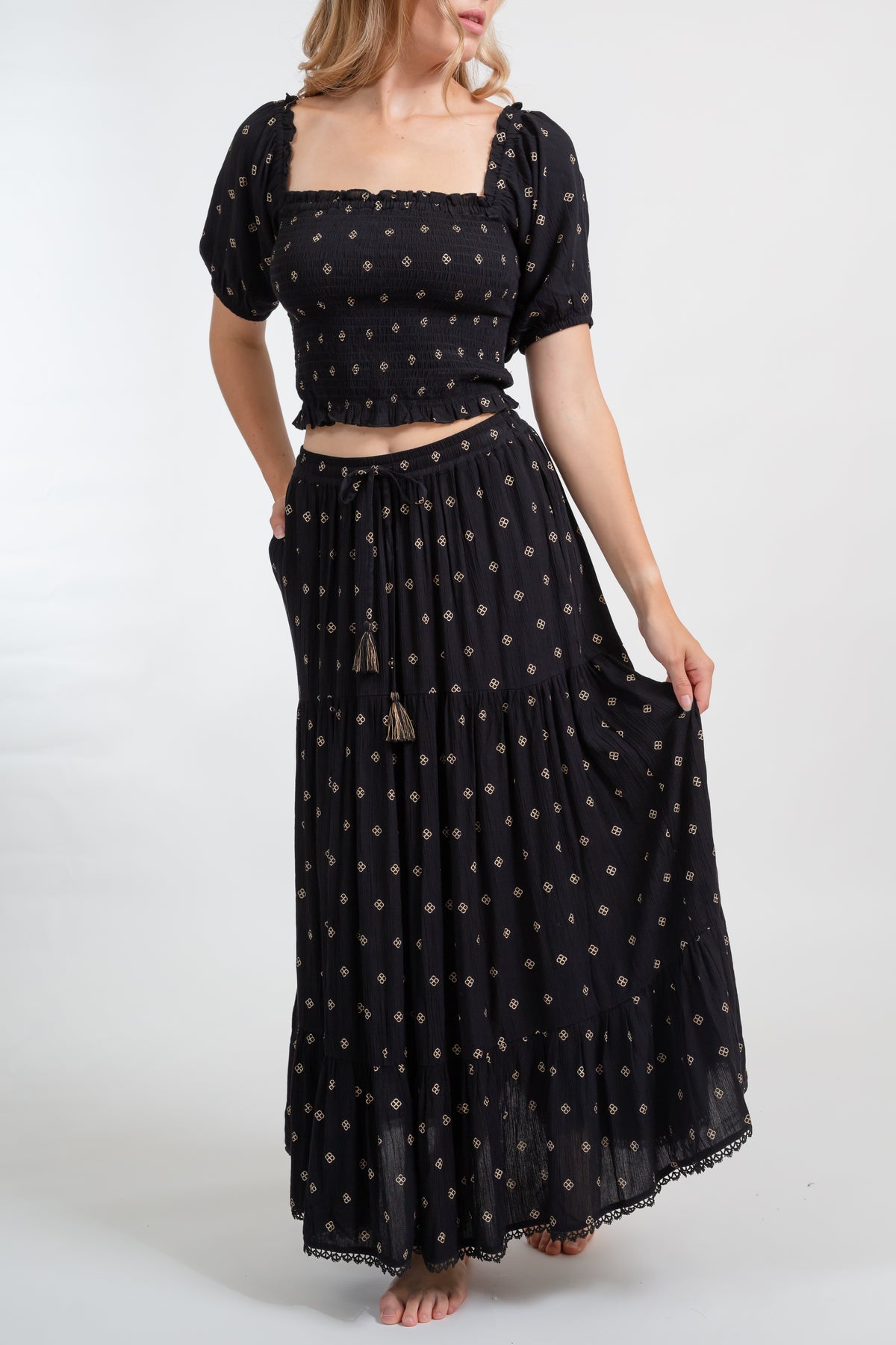a front shot of a woman model wearing a matching two piece black with gold dot print crop top and midi dress set with one hand holding the edge of her flowy dress