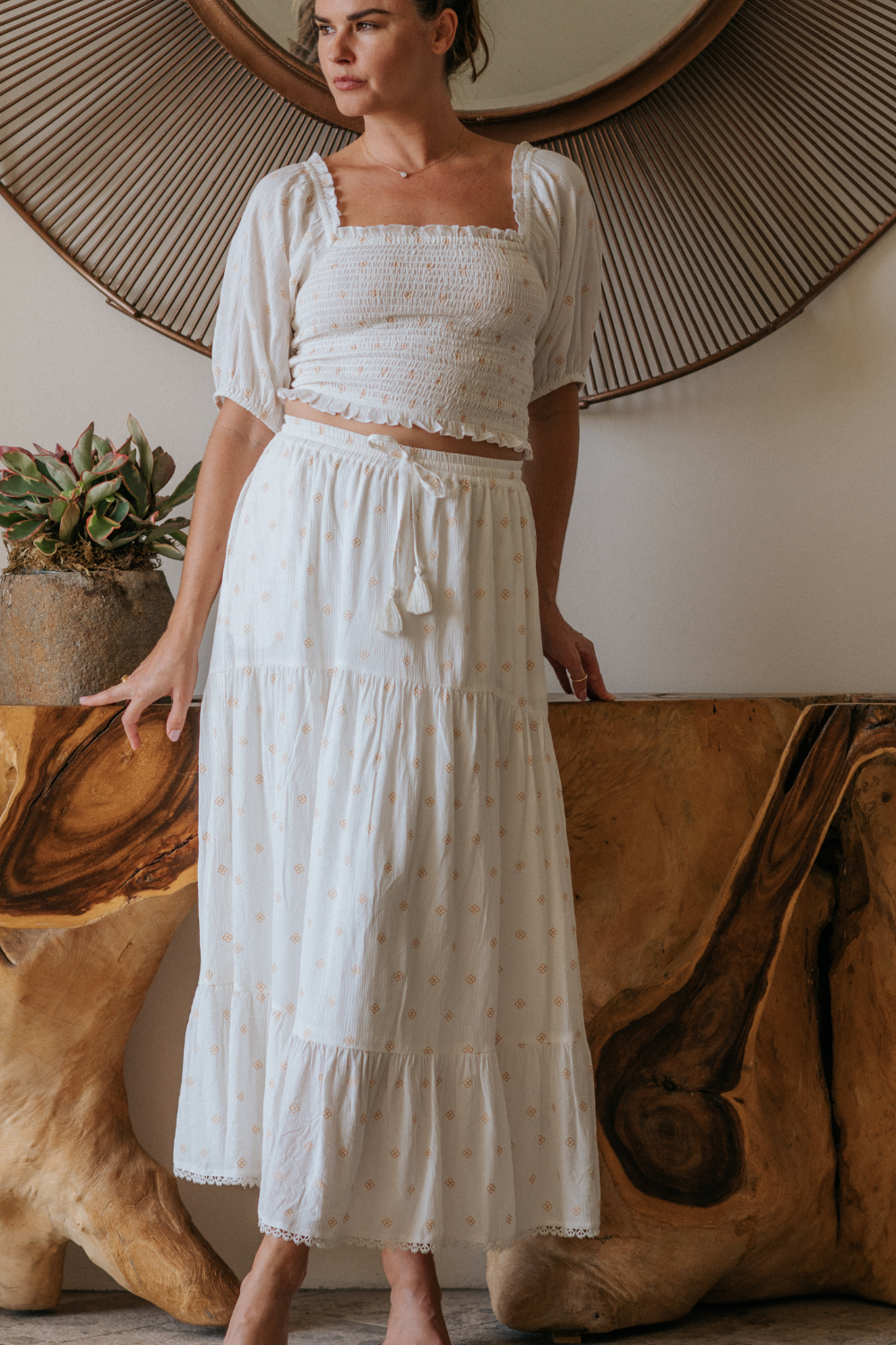 a woman model standing in front of european aesthetic decorations wearing a two piece white with gold dot print crop top and tiered maxi skirt from Koy Resort