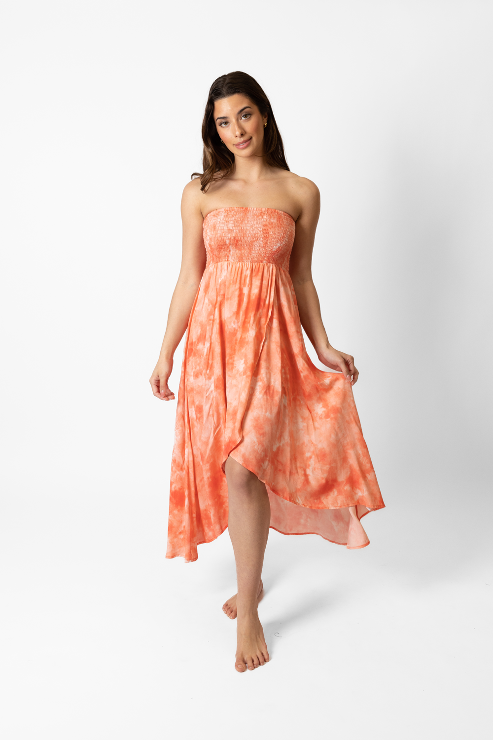 A woman standing in a photo shoot studio, wearing a coral tie-dye dress. The dress is a vibrant shade of pink, with a unique tie-dye pattern that creates a marbled effect. It has a flowing, loose fit that skims the top of her knees.