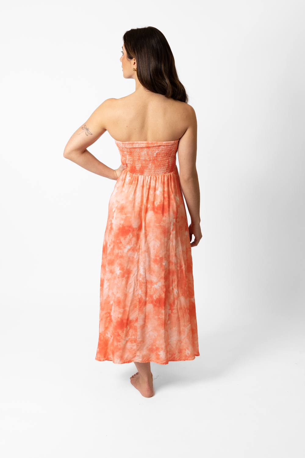 The back of a woman with one hand on the waist and the other relaxed wearing Koy Resort Aquarelle spring 2023 collection coral tie-dye dress that drops to her ankles.