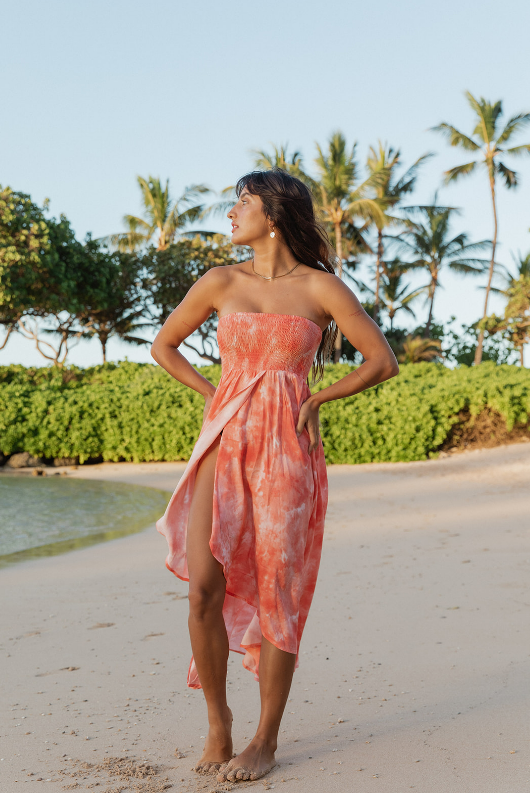 A woman standing on the beach of Hawaii wearing a coral tie-dye bandeau dress from Koy Resort