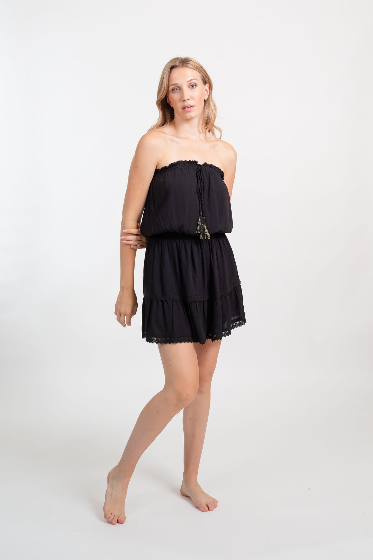 A blonde Caucasian model wearing a black color bandeau  mini boho dress, standing in a studio with her arms behind her back looking at the camera posing.