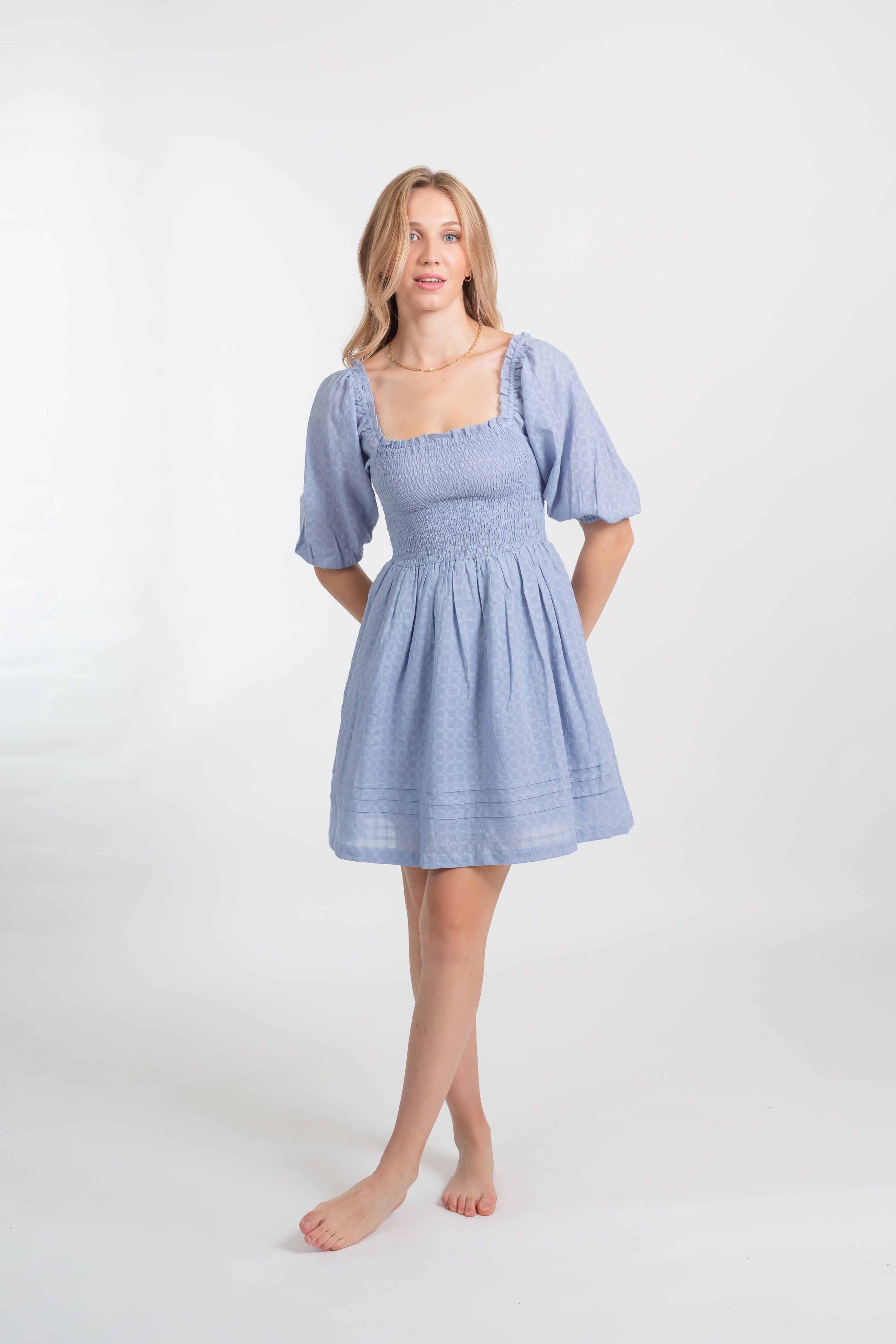 a blonde female model wearing a sky blue smocked short sleeve mini dress from Koy Resort facing forward with her hands behind her back