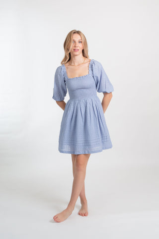 a blonde female model wearing a sky blue smocked short sleeve mini dress from Koy Resort facing forward with her hands behind her back