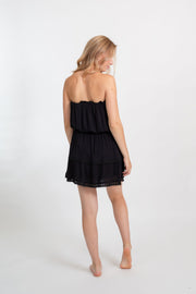 A blonde Caucasian model wearing a black  bandeau  mini boho dress, standing in a studio with her back facing and head looking over shoulder. 