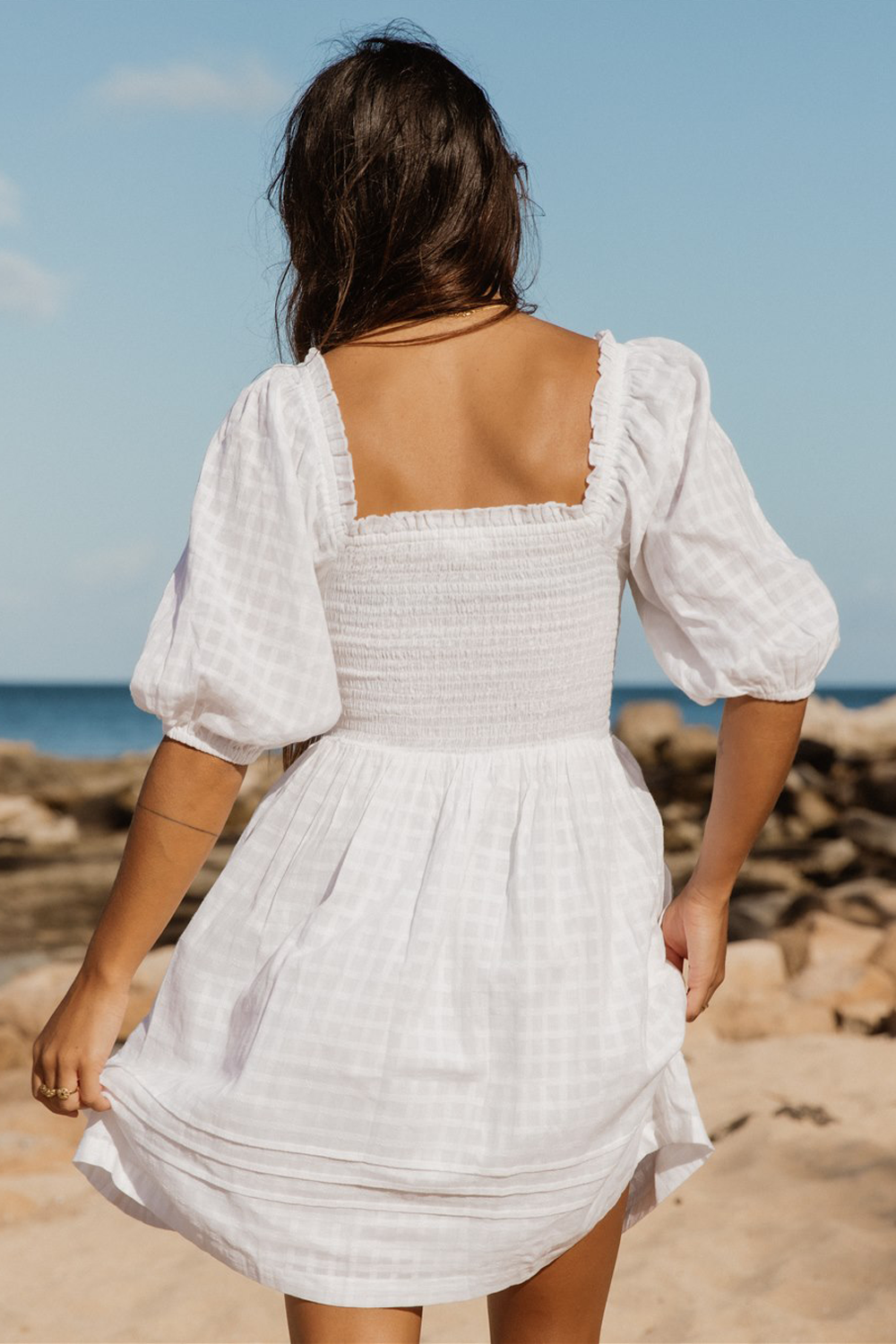 the back of a dark hair female wearing a squareed back smocked flowy mini dress standing on the beach facing the ocean