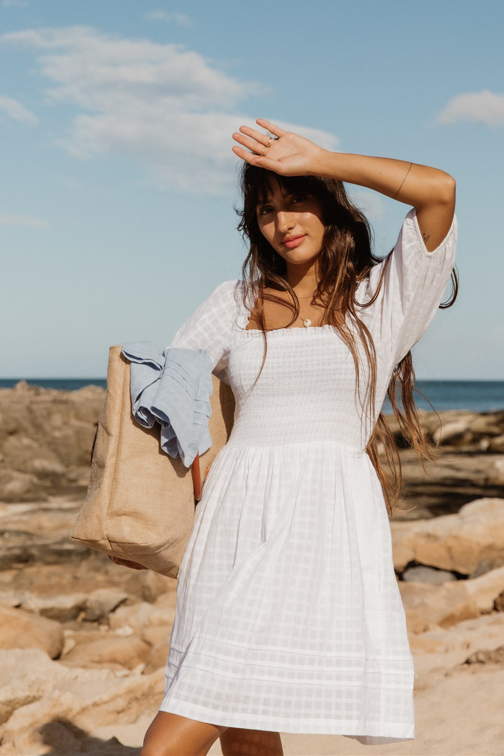 a woman wearing a white tiered smocked mini short sleeve dress from Koy Resort carrying a beach bag on the beach with one hand on her head to block the sunshine in her eyes