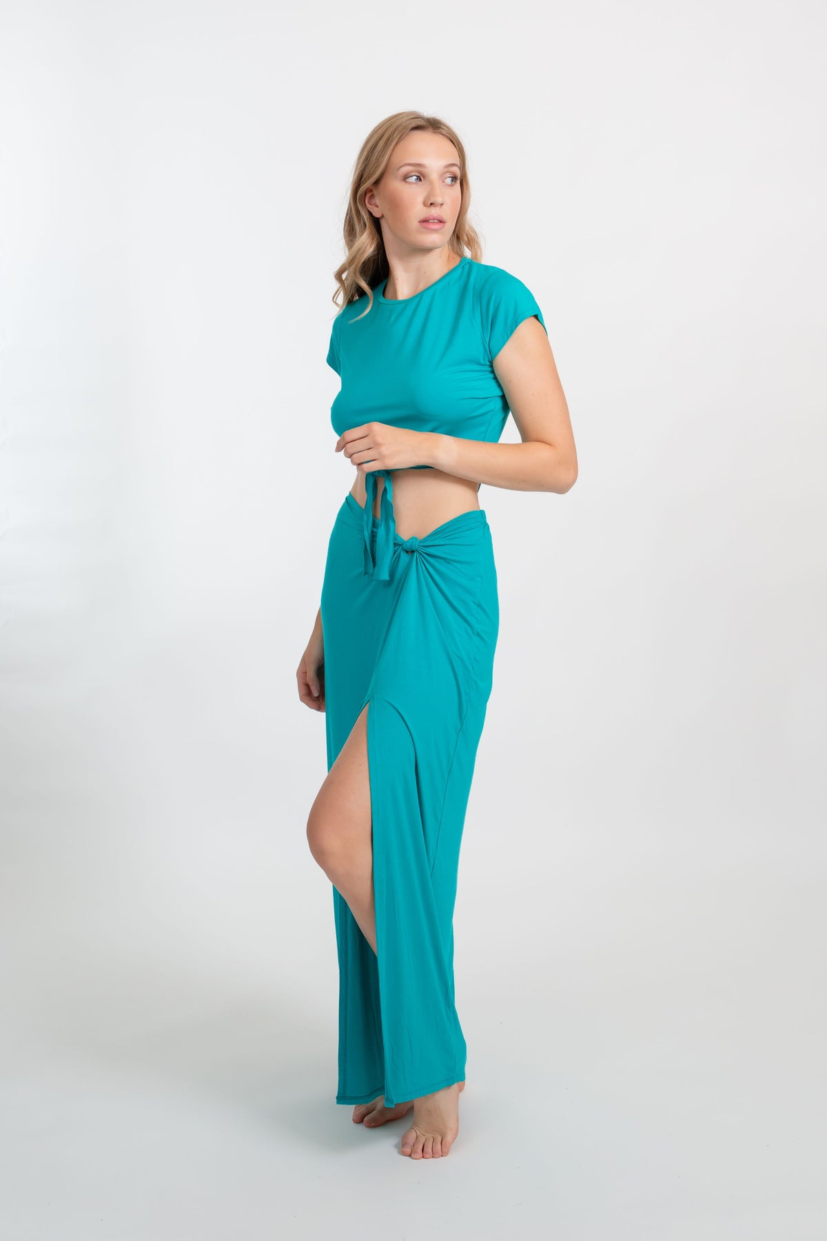 a blonde female model standing on the side wearing an aquamarine color crop short sleeves top and a matching maxi side slit maxi skirt from Koy Resort