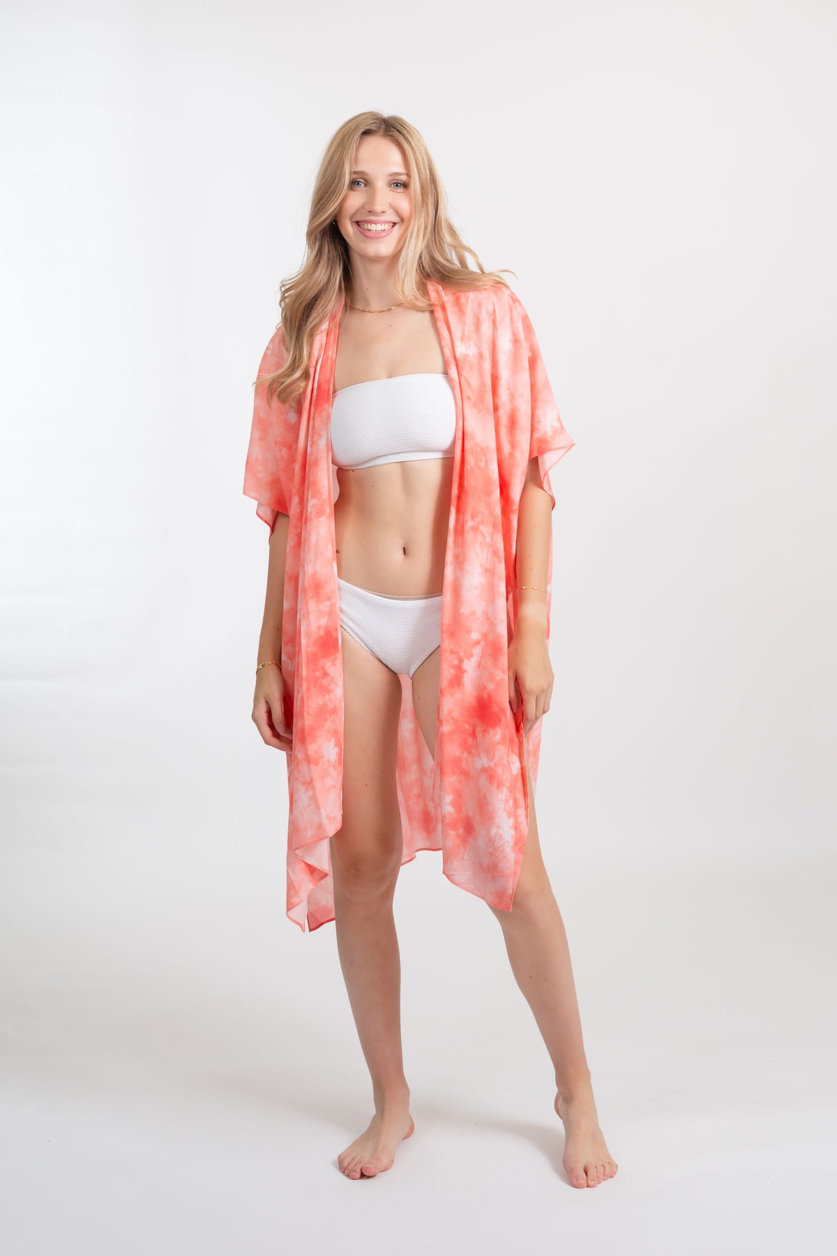 A woman wearing white bandeau two peice white swimsuit with a coral tie-dye print kimono style beach cover up smiling very happy at the camera