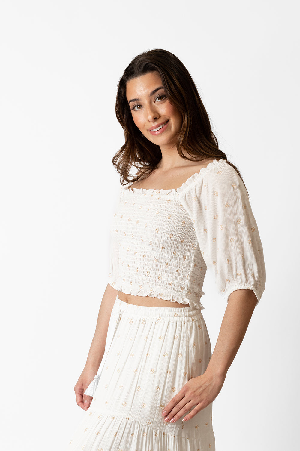 wo zoomed in side shot of a dark hair woman model wearing a white with gold dot print smocked crop top with matching skirt with one hand in her skirt pocket