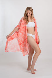 a woman wearing white bandeau two-piece swimsuit and a coral tie-dye print kimono style beach cover-up with one hand holding the edge of the kinomo