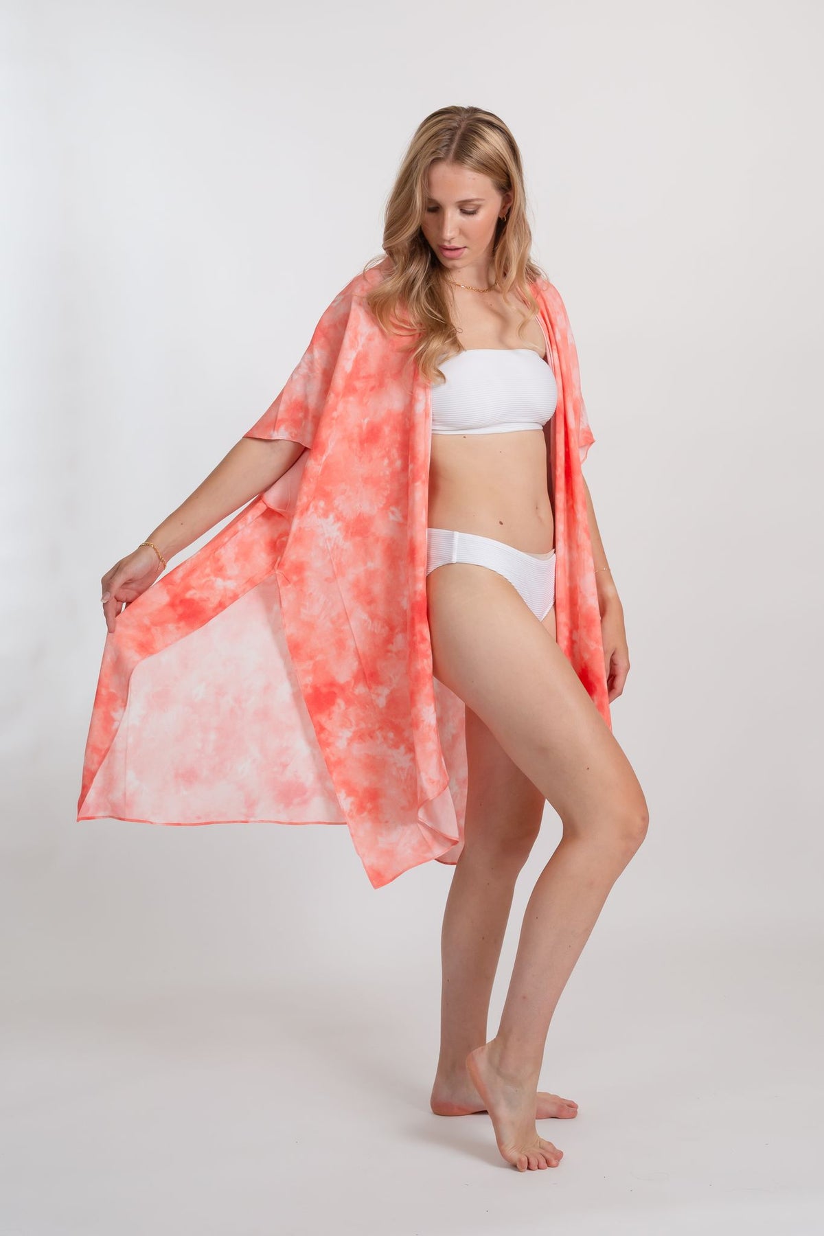 a woman wearing white bandeau two-piece swimsuit and a coral tie-dye print kimono style beach cover-up with one hand holding the edge of the kinomo 